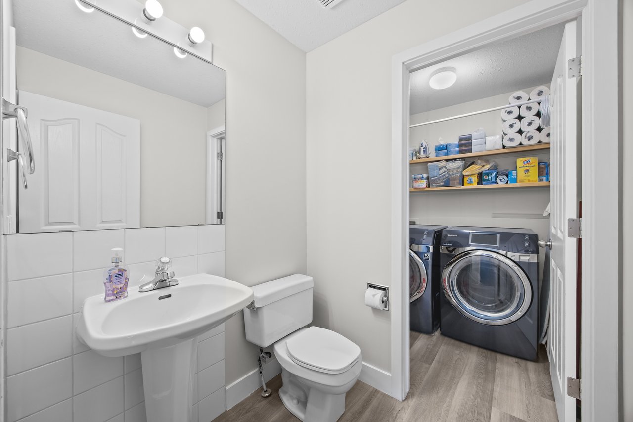 Bathroom with a pedestal sink and toilet opening to a stocked laundry area with front-loading machines.