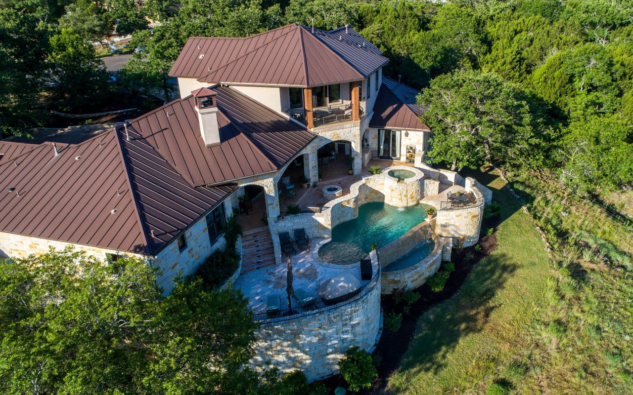 Aerial view of a stone house with a brown metal roof and a curved pool terrace.