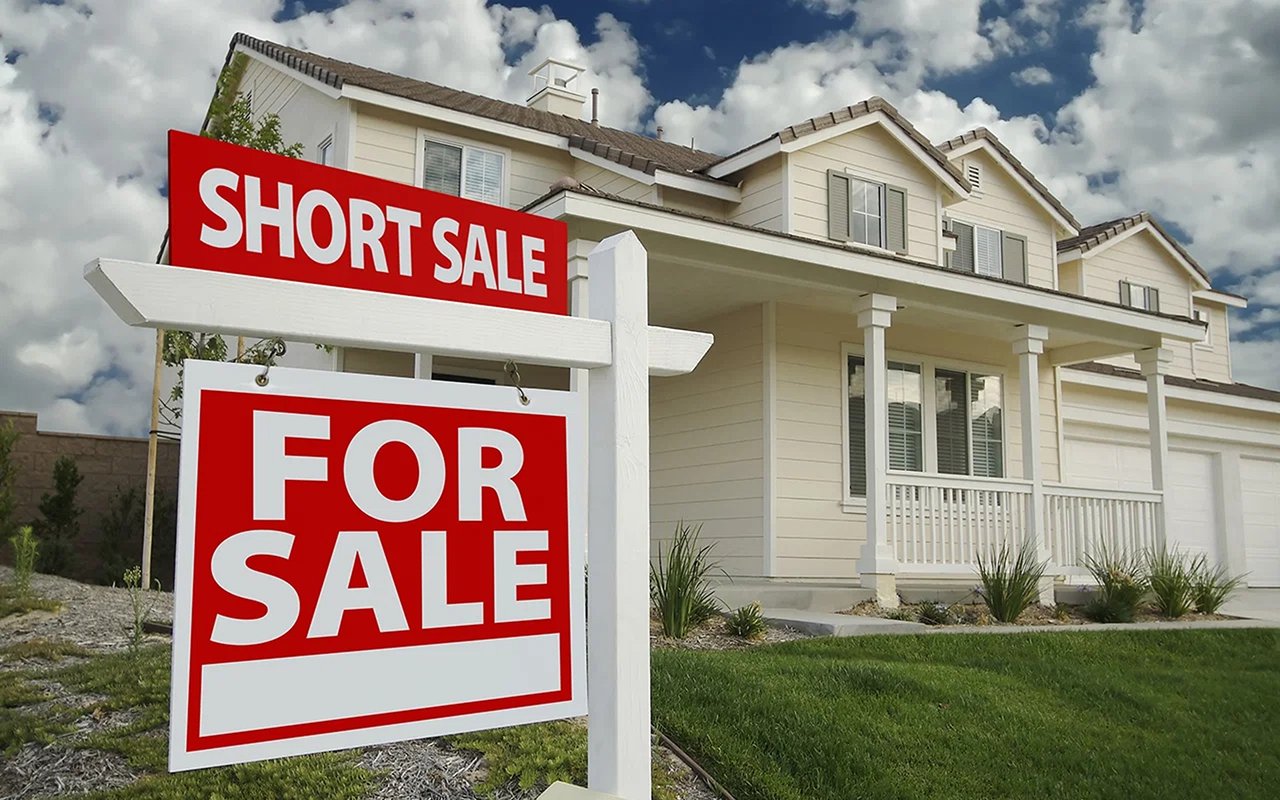 Two-story cream house behind a prominent red short sale and for sale sign.