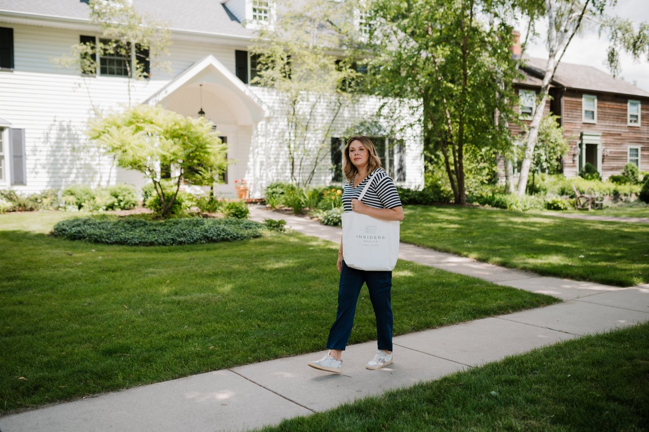 Cait Berry of Insiders Realty walking along a sidewalk in front of a Madison-area home, carrying an Insiders Realty tote, representing the kind of in-person neighborhood evaluation that helps buyers understand what a home actually feels like in context.