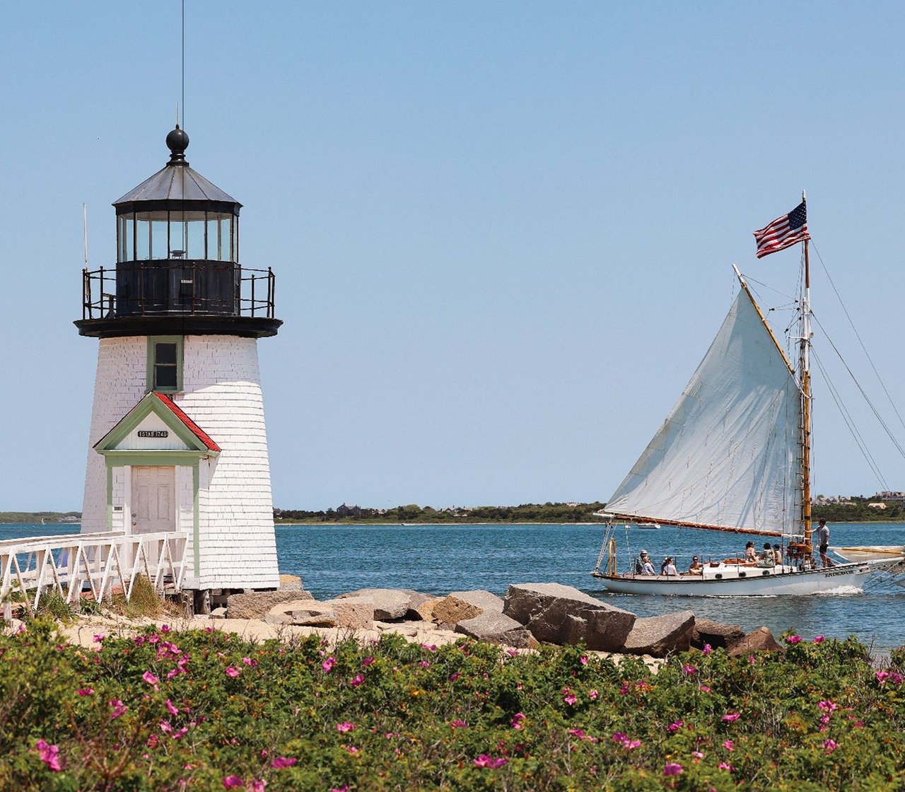 White lighthouse beside the water as a sailboat with an American flag passes.
