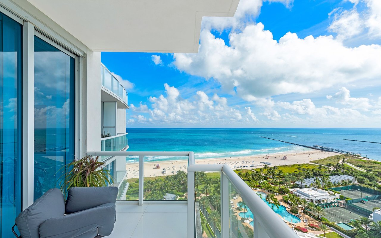 High-rise balcony overlooking a beach and turquoise ocean, with glass railings and a gray lounge chair.