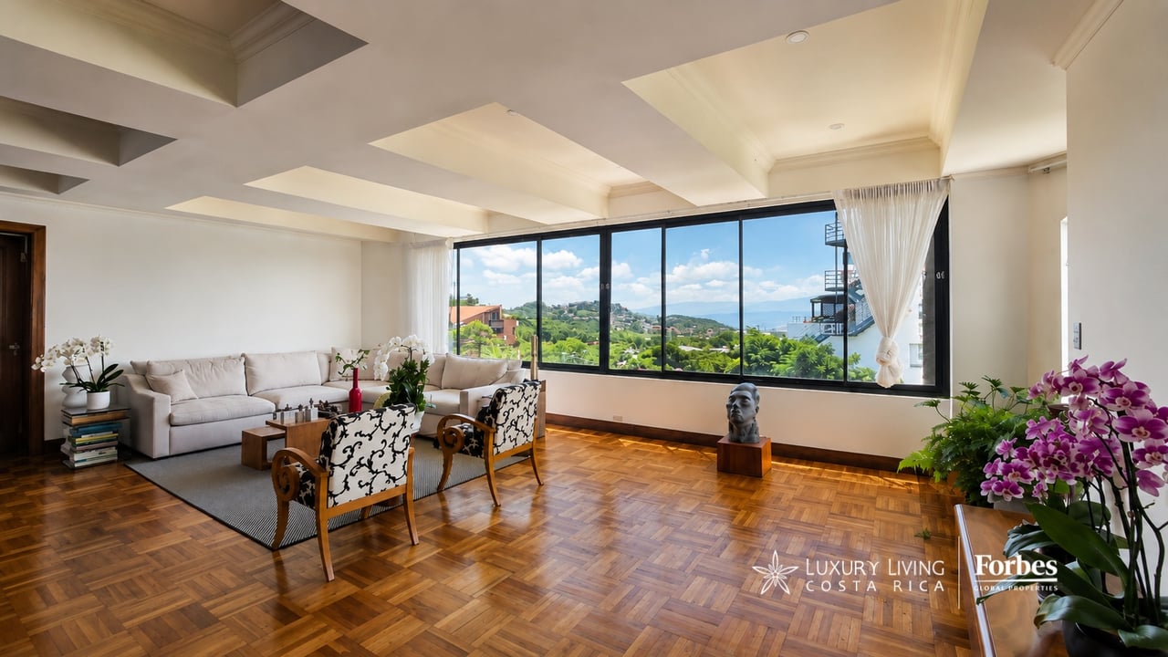 Condominium living room with parquet floors and wide windows overlooking green hills and distant mountains.