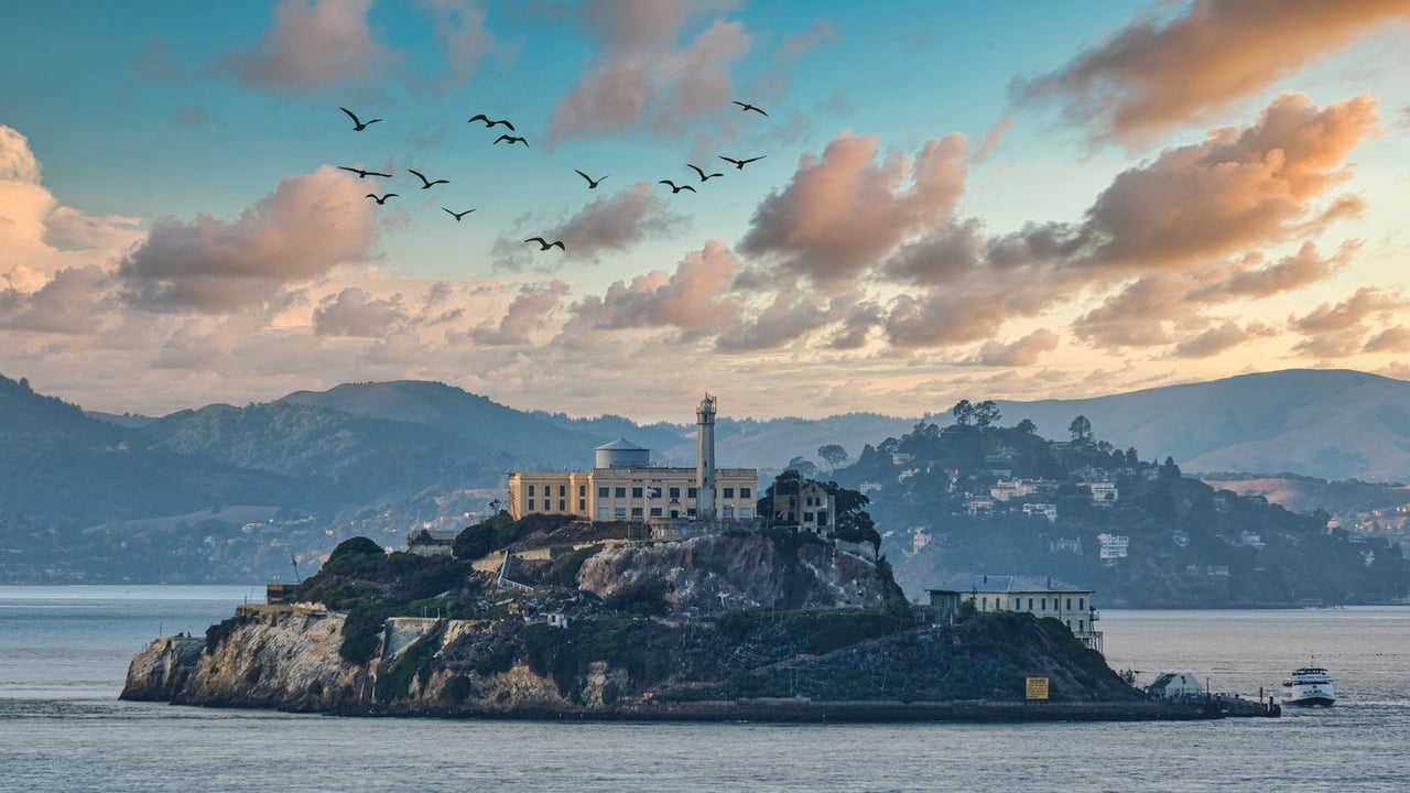 alt="Color photograph of Alcatraz Island in San Francisco Bay during sunset, showing the rocky island with the prison buildings on top, a lighthouse, and a water tower, surrounded by water with birds flying in the sky, distant hills, and a small boat nearby under a pastel-colored, cloudy sky."