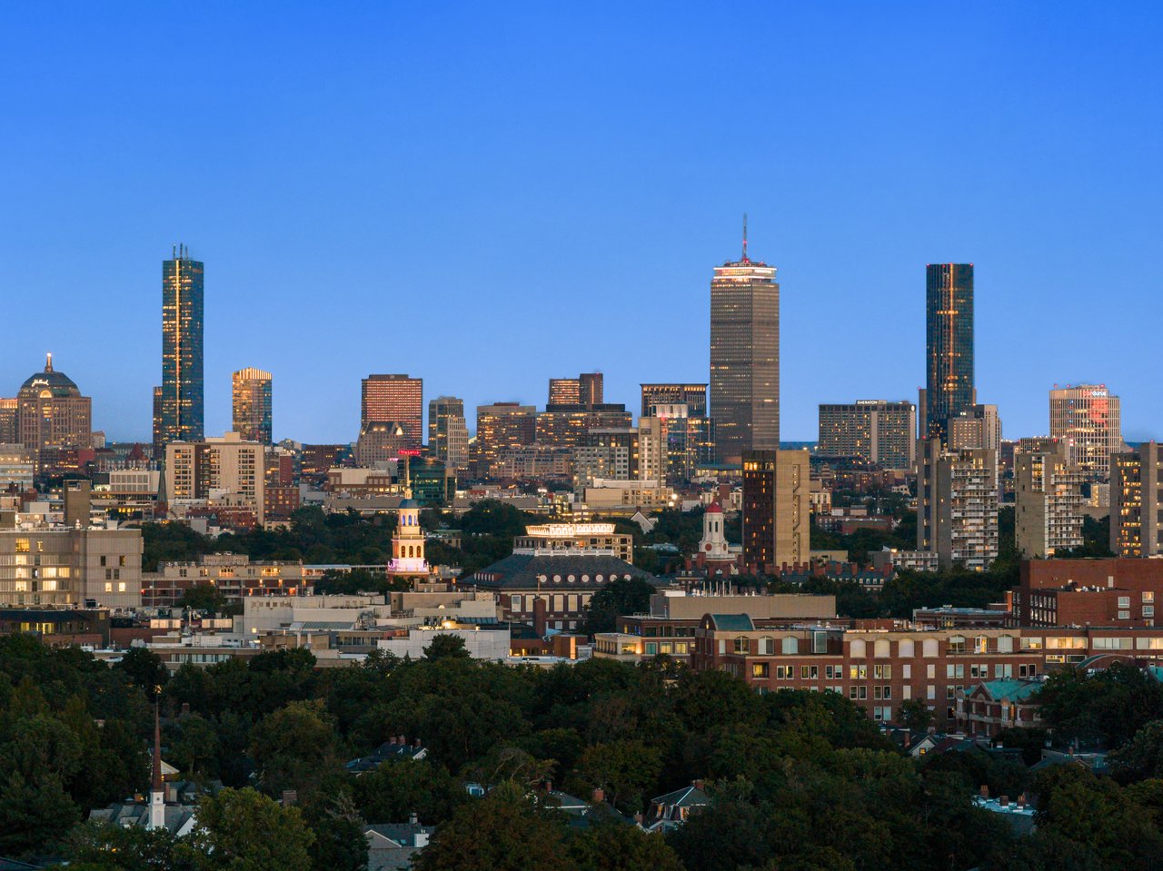 Boston skyline at dusk above tree-lined neighborhoods.