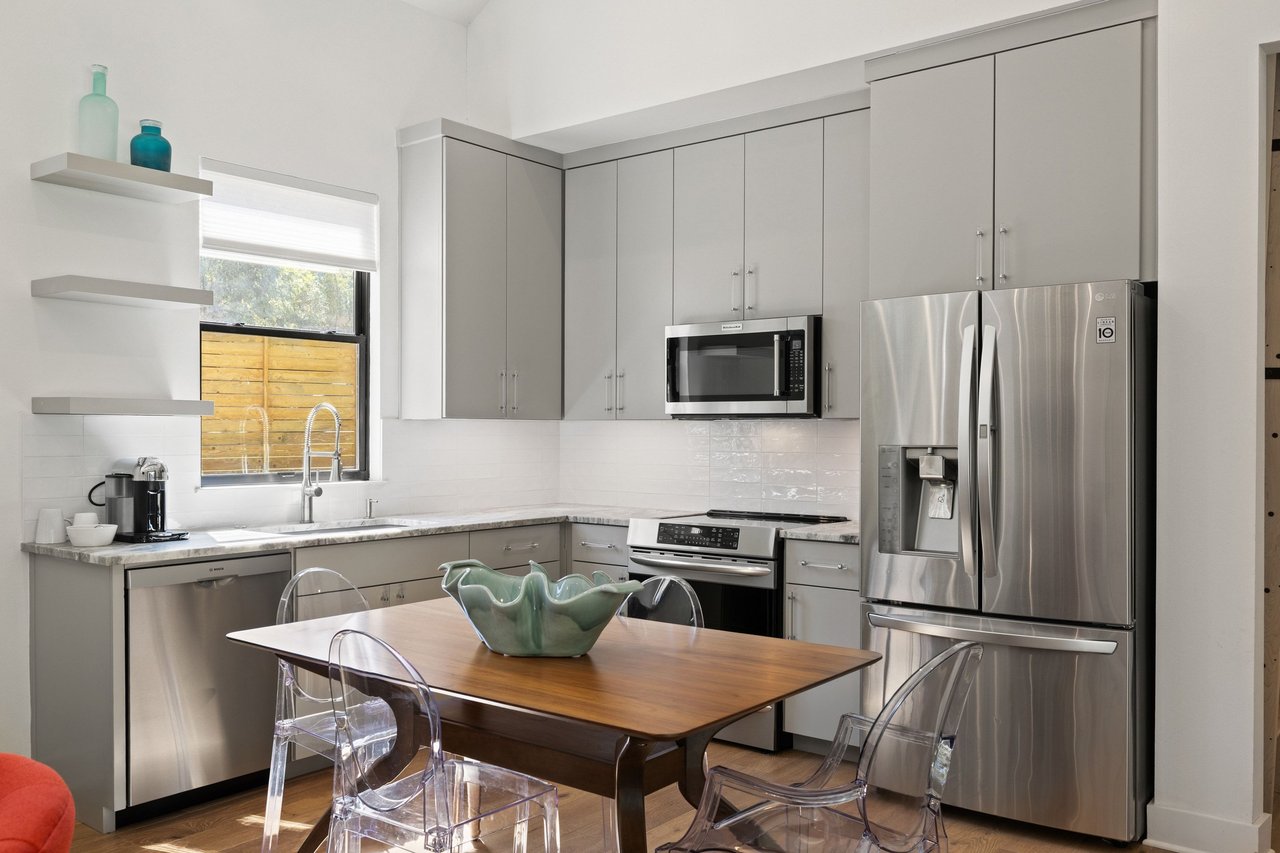 Modern kitchen with gray cabinets, stainless steel appliances, and a wood dining table with clear chairs.