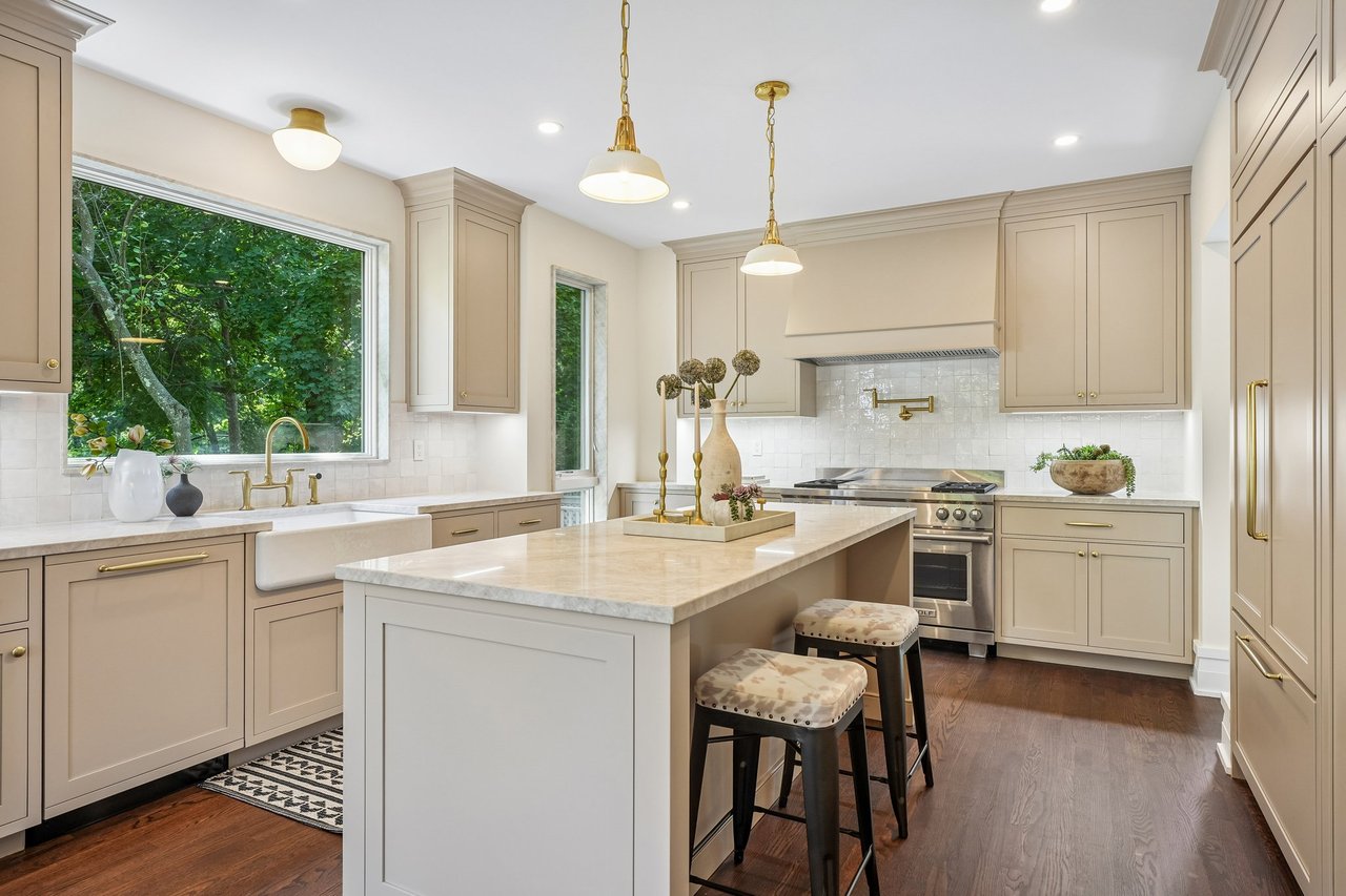 Bright kitchen with cream cabinets, marble island, farmhouse sink, and leafy views through a large window.