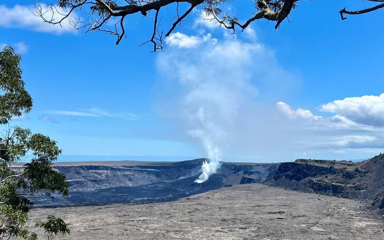 Steam rises from a volcanic crater, framed by leafy branches beneath a bright blue sky.