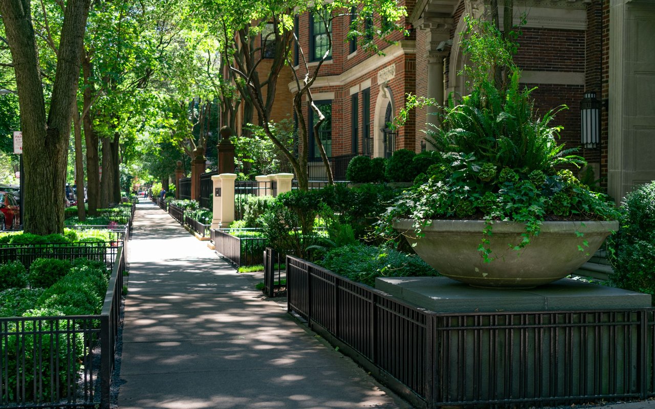 Tree-lined residential sidewalk beside brick homes, with iron fences and a large stone planter.