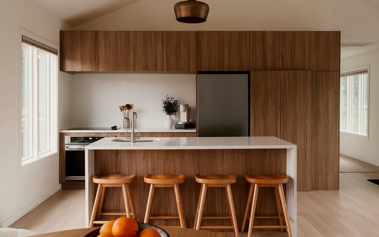Modern kitchen with walnut cabinetry, a white island, four wooden stools, and large windows.
