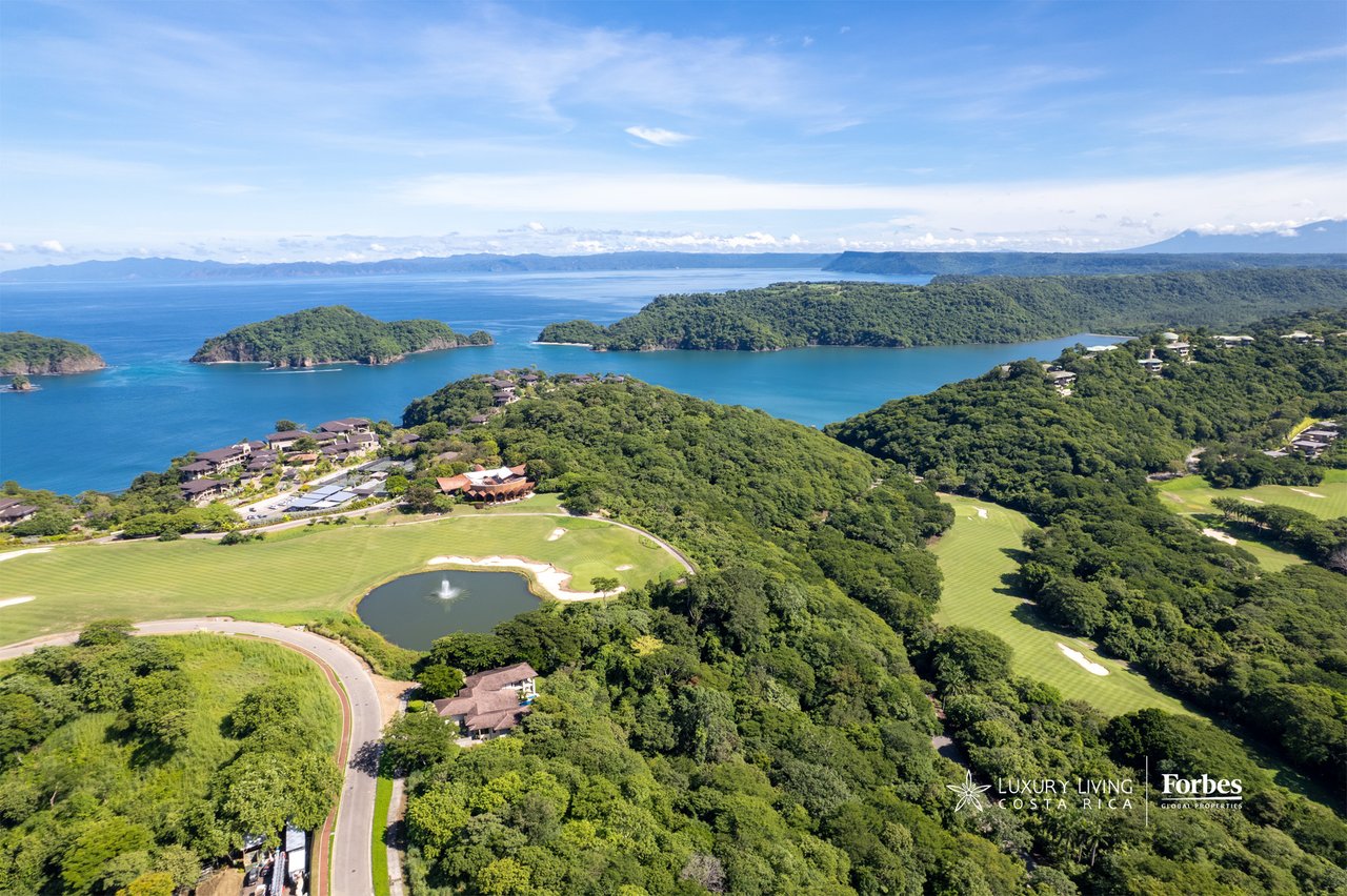 Aerial view of coastal homes and golf fairways amid green hills beside blue ocean inlets.
