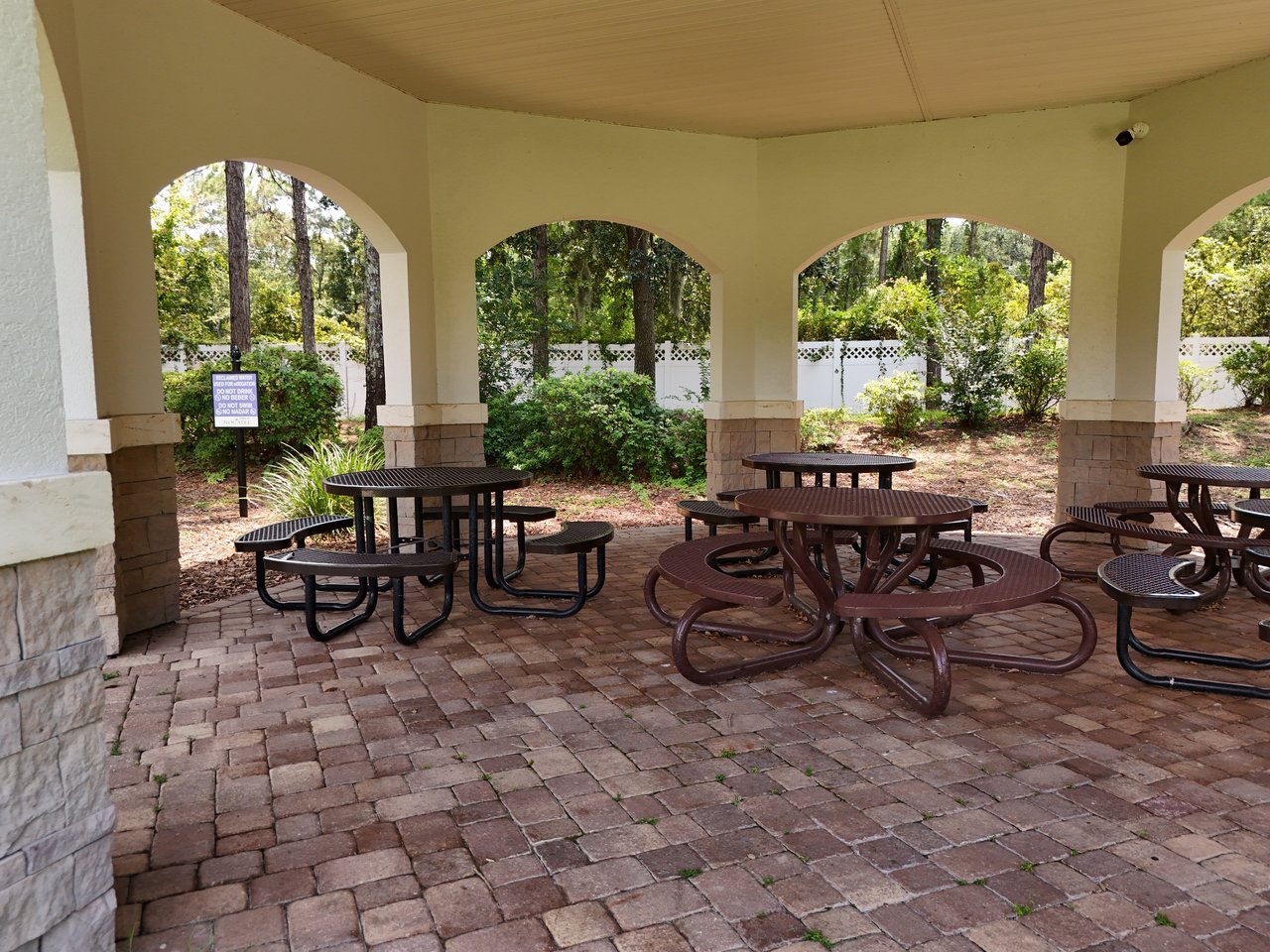 Covered community pavilion with arched openings, brick paving, and picnic tables among trees.