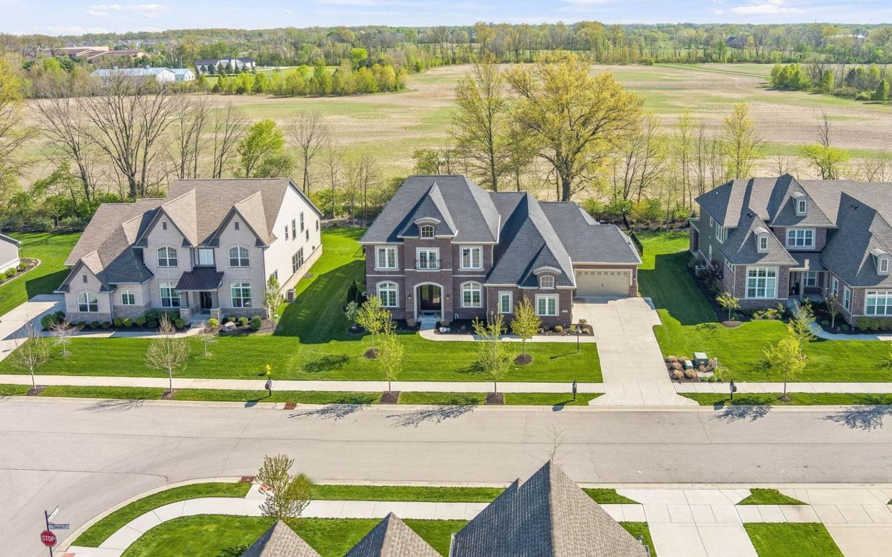 An elevated, high-angle aerial view showcasing three large, two-story luxury homes with multi-gabled roofs built next to each other along a suburban street. Perfectly manicured green lawns and sidewalks separate the properties from the road, while a vast open green field stretches out directly behind the houses under a clear sky.