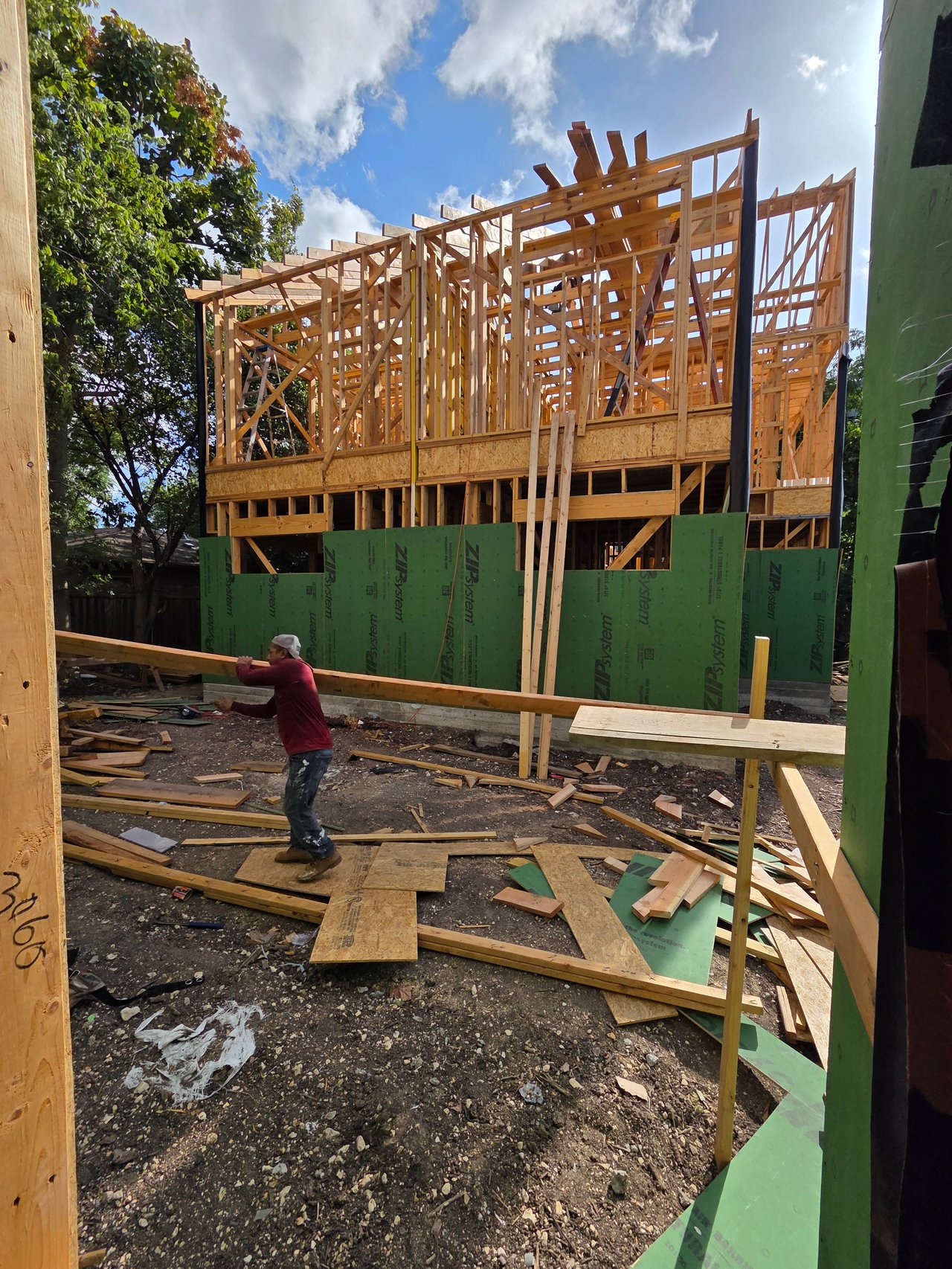House under construction with exposed framing and a worker handling lumber in the foreground.