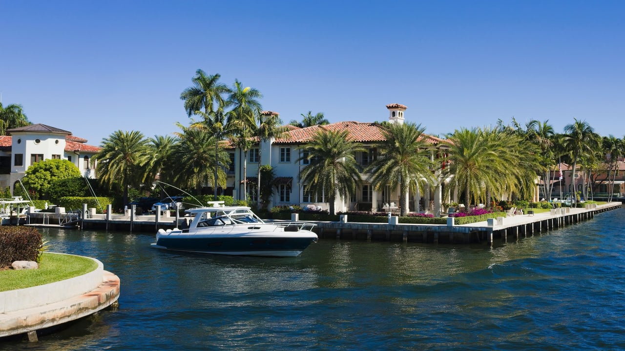 Mediterranean-style waterfront estate with terracotta roofs, palm trees, and a private dock with a moored boat.
