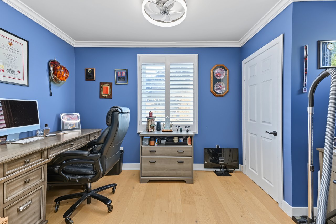 Blue home office with a wood desk, black chair, and window with white plantation shutters.