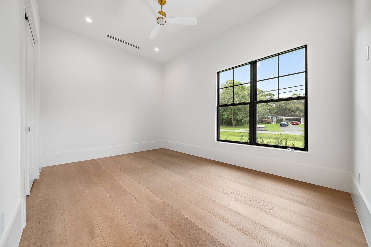 Empty bedroom with light wood floors, a ceiling fan, and a large window overlooking greenery.