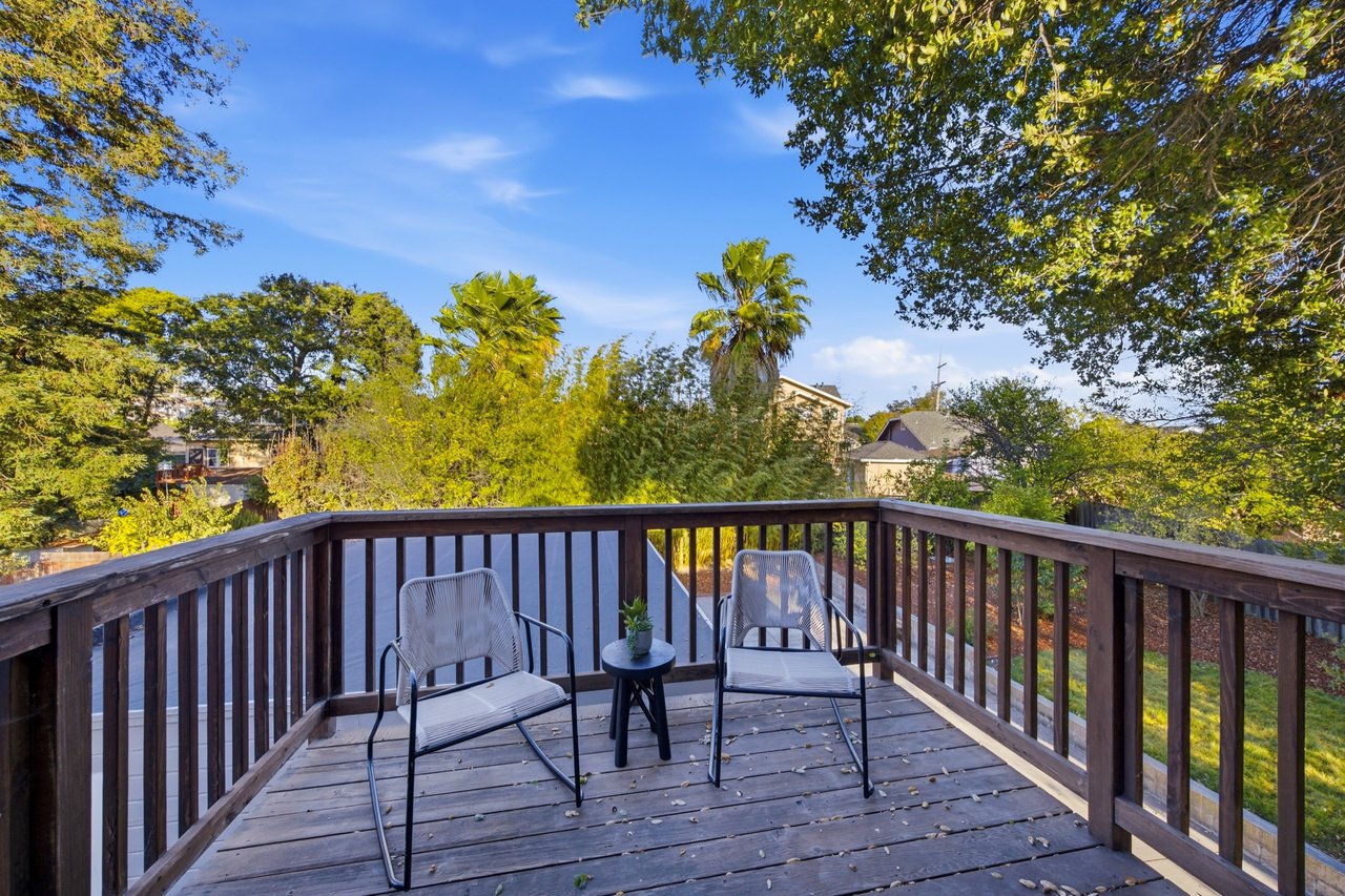 Wooden deck with two chairs and a small table, overlooking leafy trees and nearby homes.