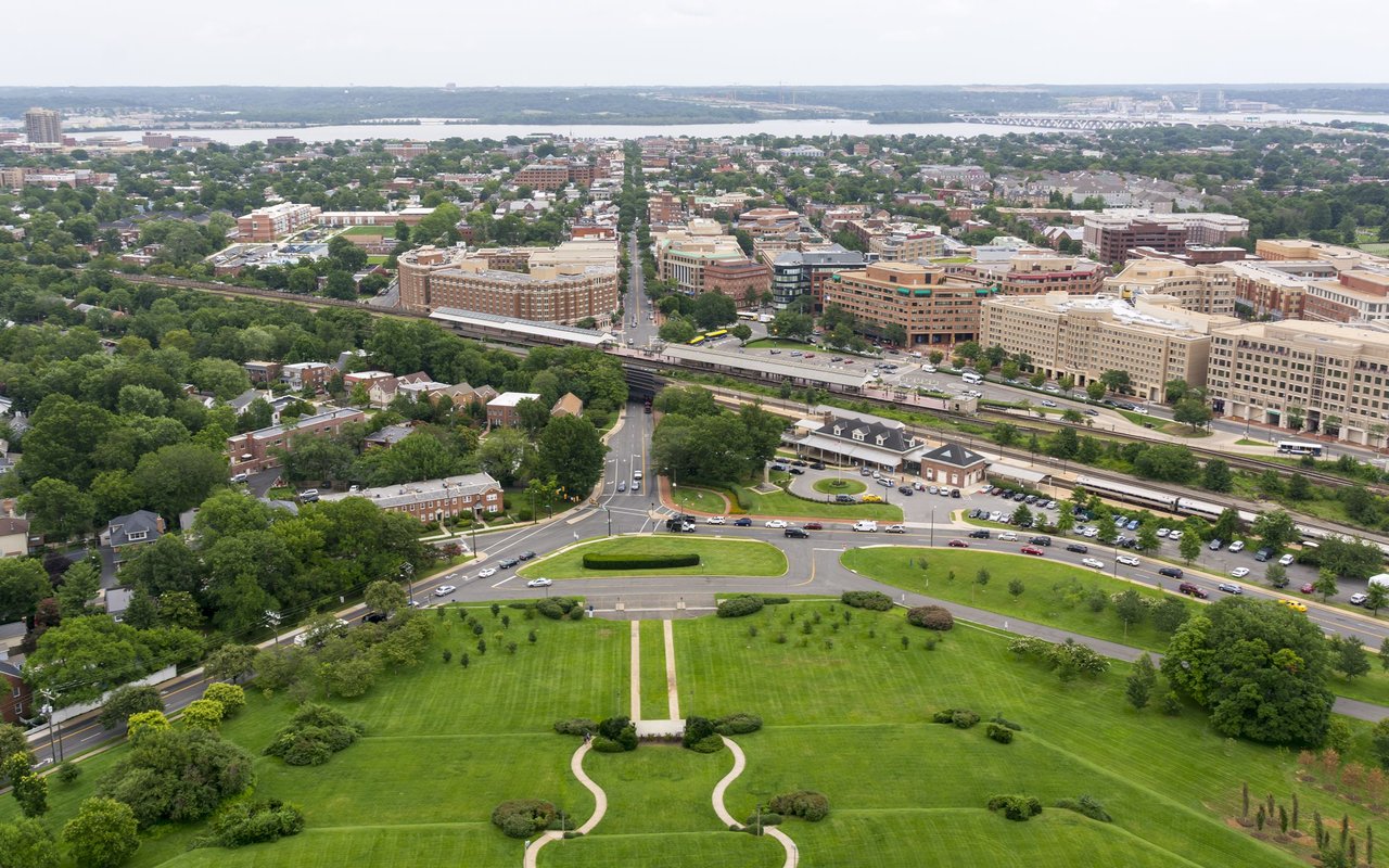 Panoramic aerial view of Alexandria, with green park grounds, downtown buildings, and the Potomac River in the distance.