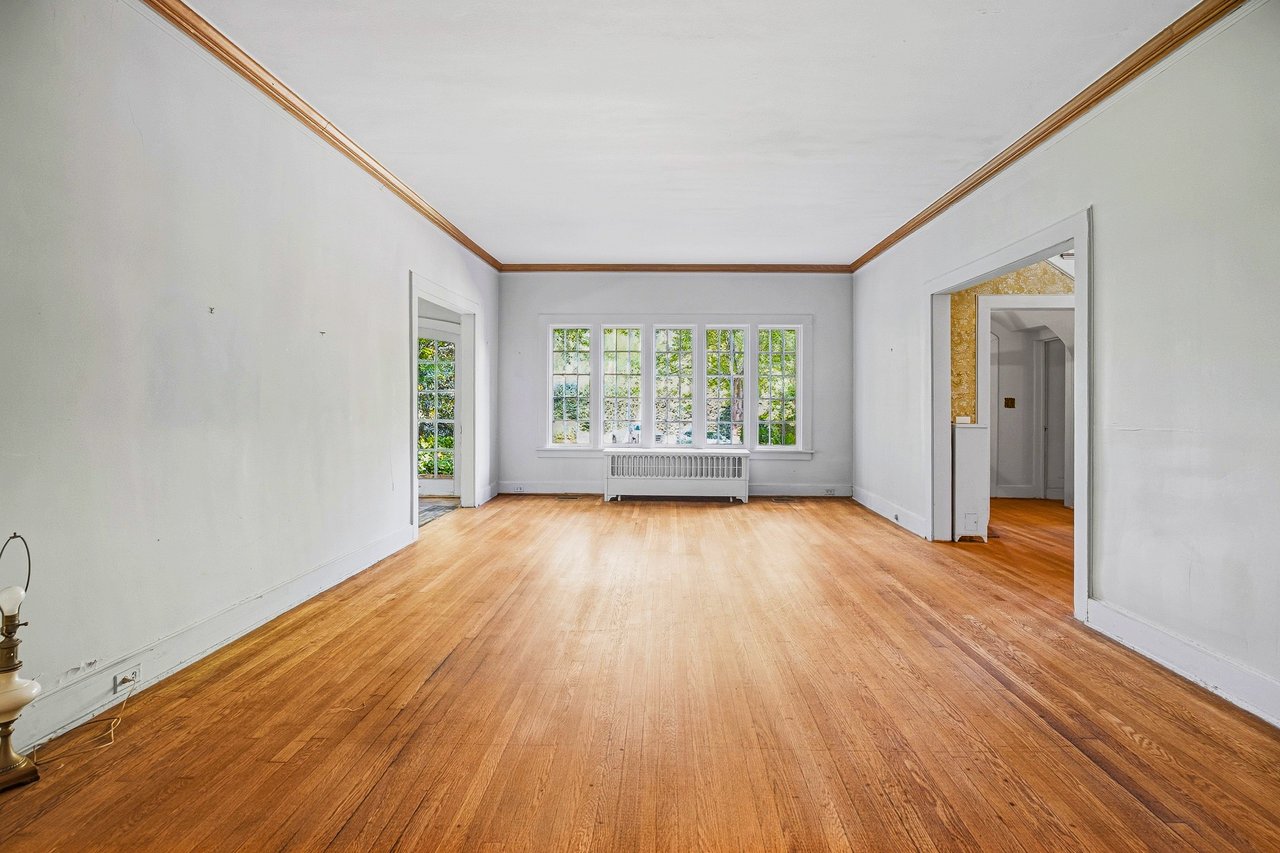 Empty living room with hardwood floors, wood crown molding, and multi-pane windows above a radiator.