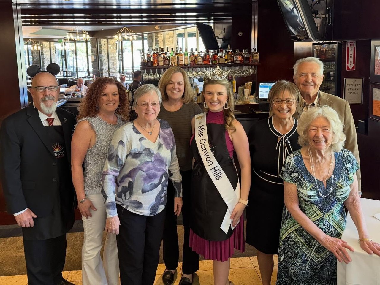 Eight adults pose in a restaurant; a crowned woman wears a Miss Canyon Hills sash.