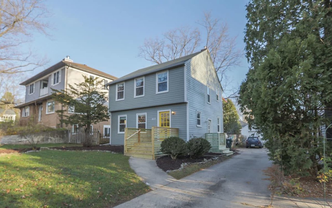 Two-story gray house with a yellow front door and a small fenced entry deck.