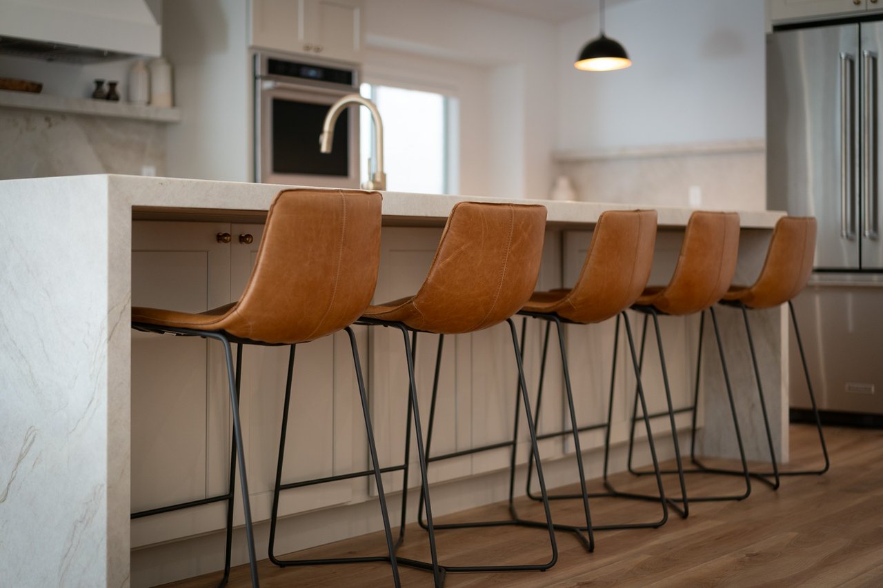 Modern kitchen with a marble island and five tan leather bar stools.