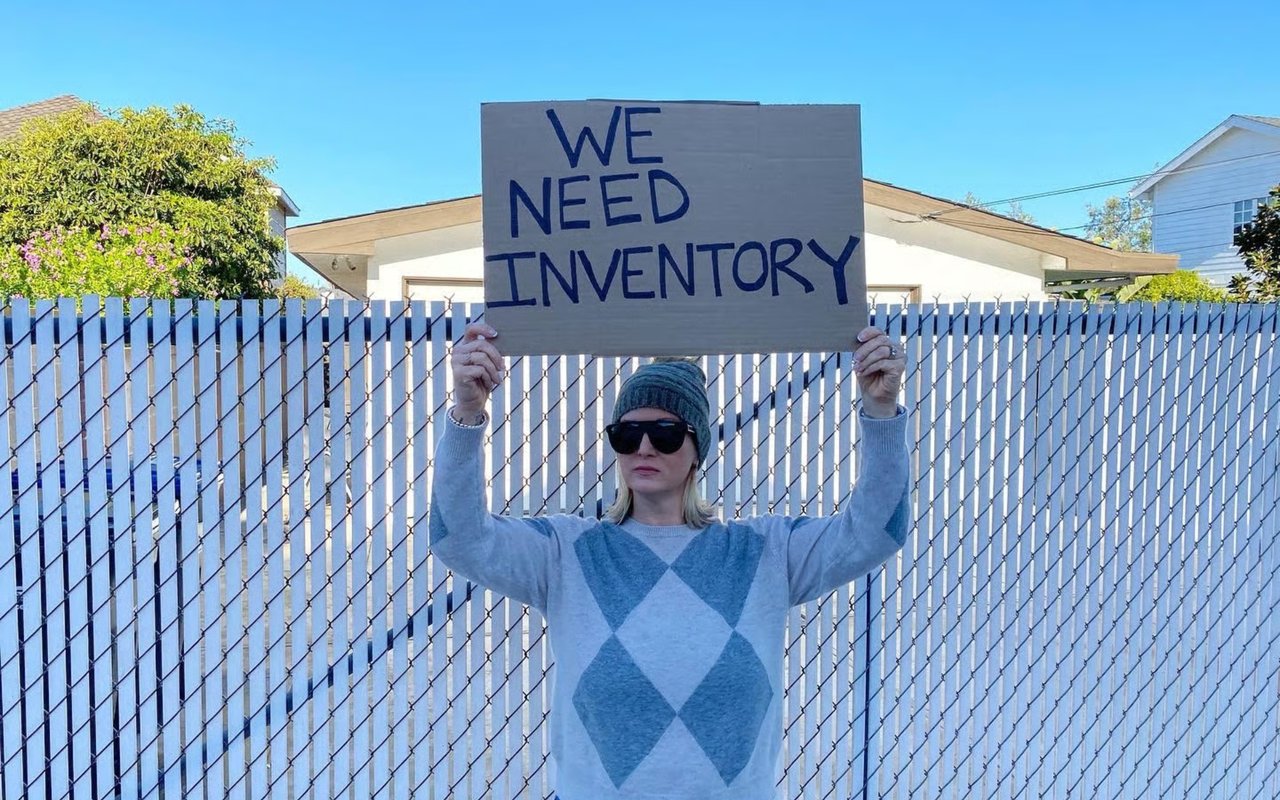 A woman holds up a cardboard sign saying "We Need Inventory" in front of a white fence and a house.