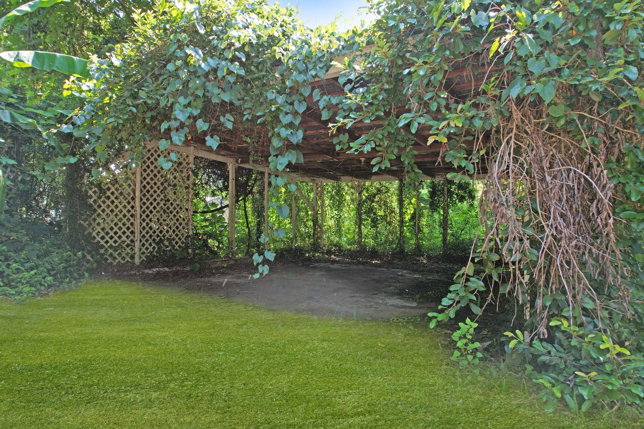 Overgrown wooden structure surrounded by lush vines and greenery, creating a secluded, natural shelter. Sunlight filters through leaves, casting shadows on grass.