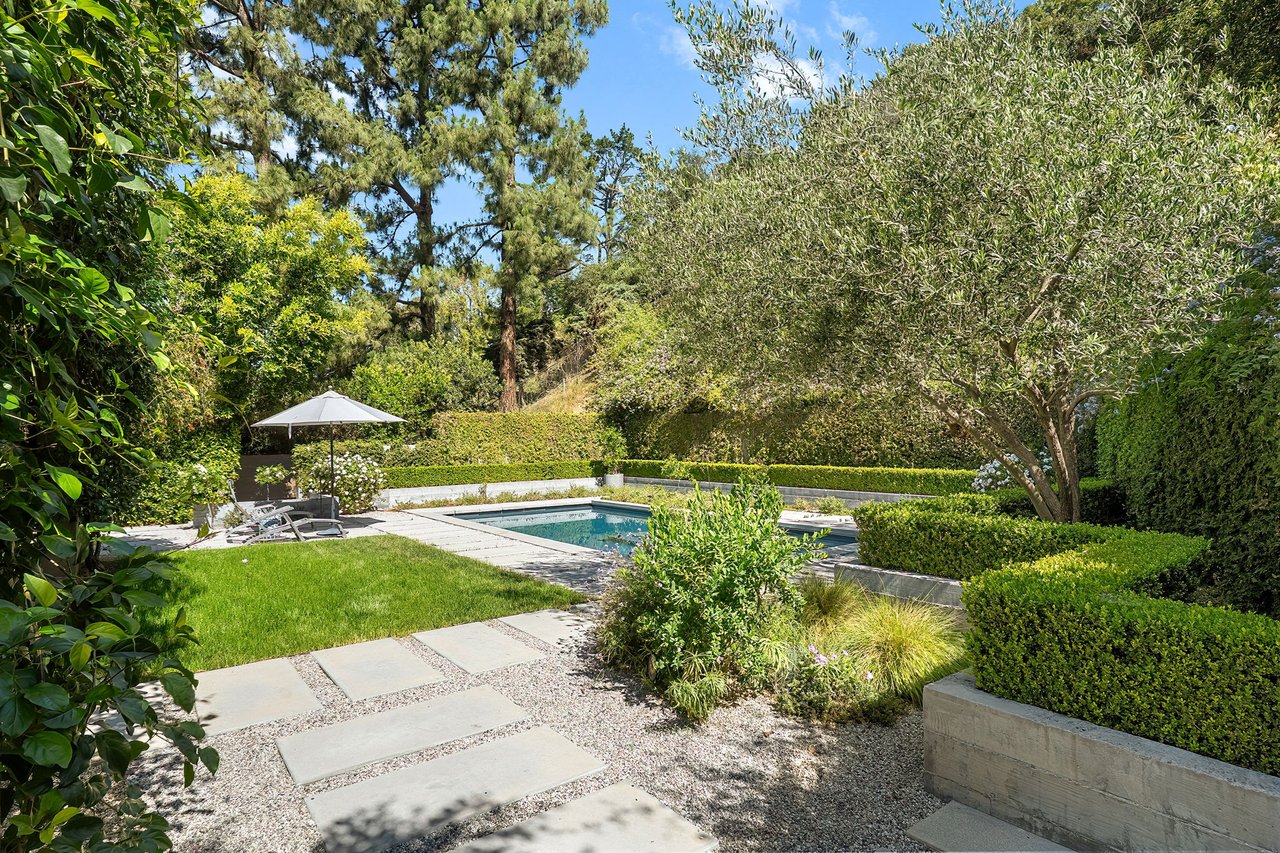 Backyard pool bordered by a stone patio, trimmed hedges, and mature trees.