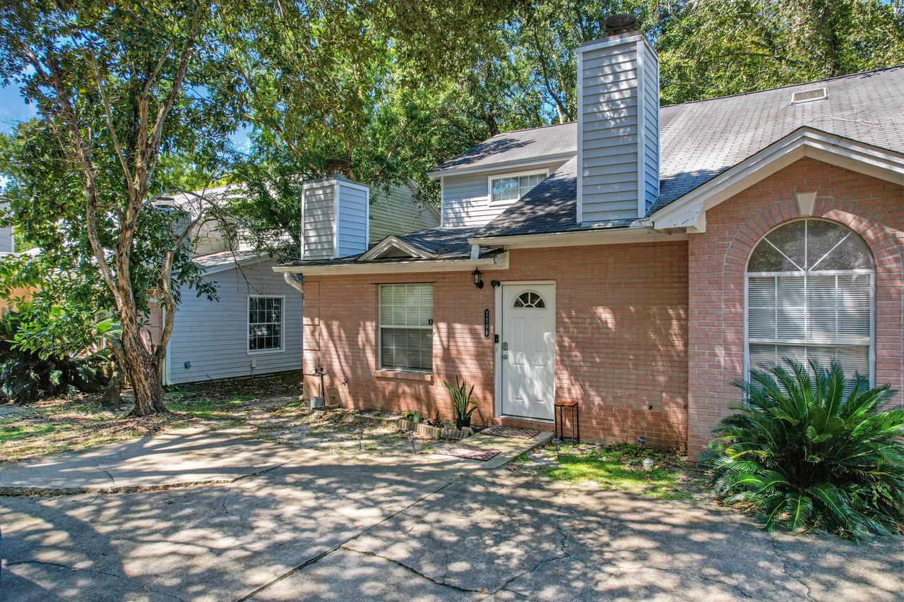Closer view of the front exterior of a brick townhome at 2506 Nugget Lane in Tallahassee, Florida. The home features a white front door, rectangular and arched windows with white trim, and two chimneys with gray siding. A concrete driveway and walkway lead to the entrance, with small plants and shrubs near the foundation.