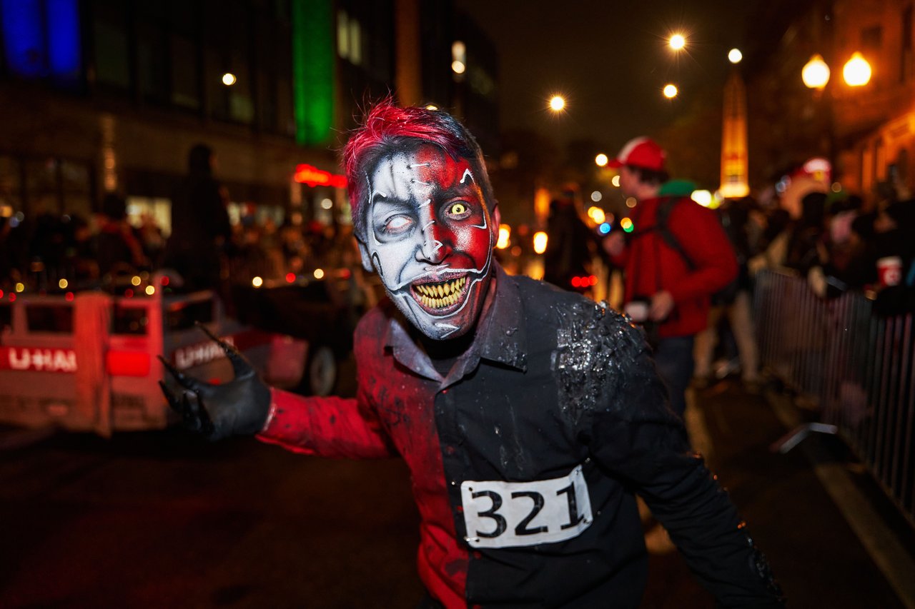A wide nighttime photograph of the NYC Village Halloween Parade on Sixth Avenue, featuring large crowds in Halloween costumes, police barricades, closed Manhattan streets, and illuminated buildings.