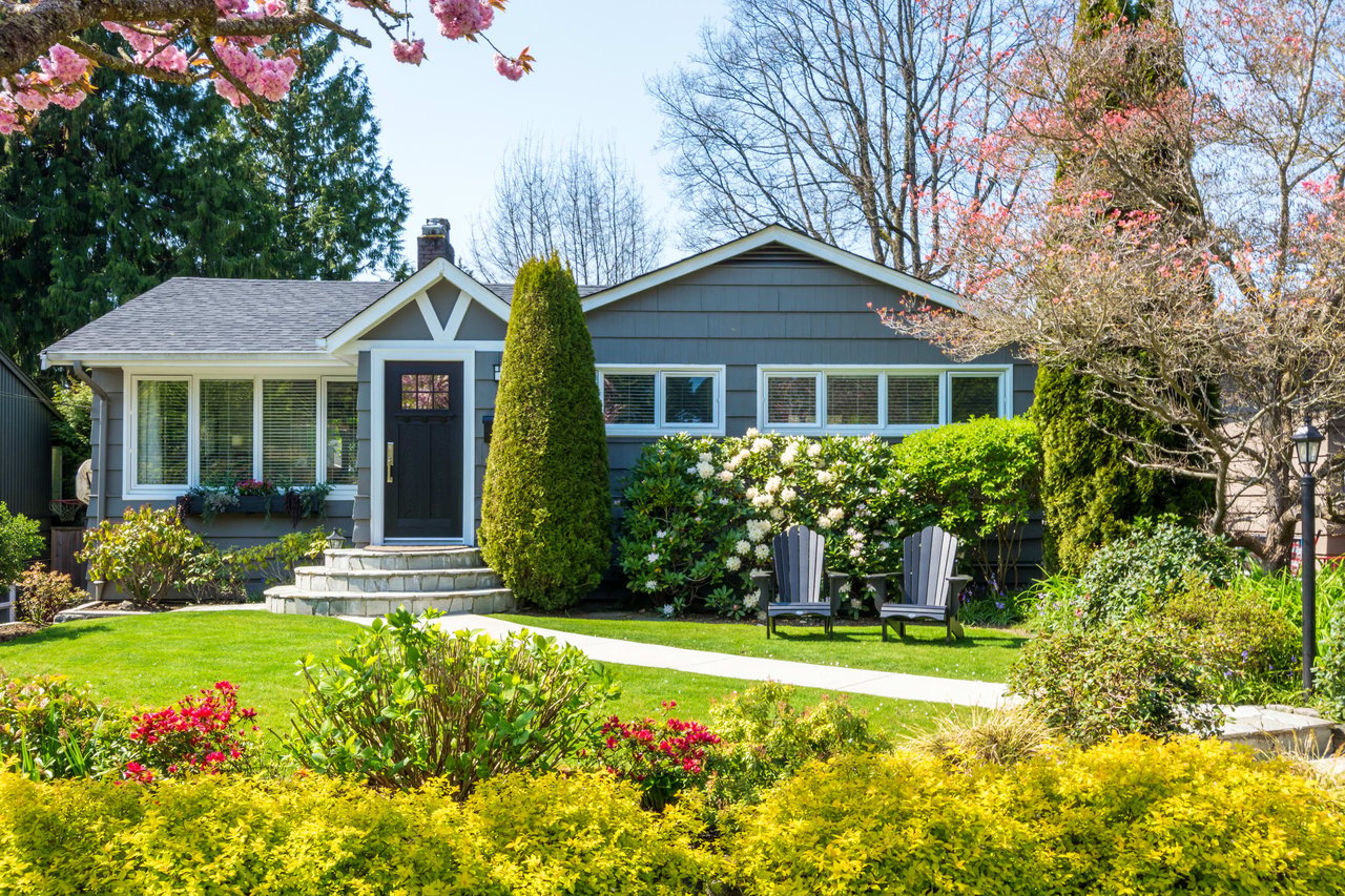 A two-story colonial-style house with a black exterior, white shutter, and a lawn with flowers in front of it.