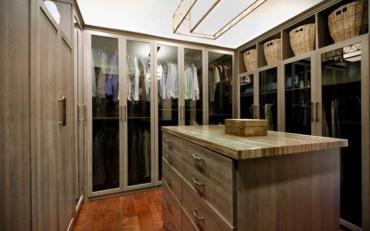Walk-in dressing room with glass-front wardrobes, woven baskets, and a large wooden island.