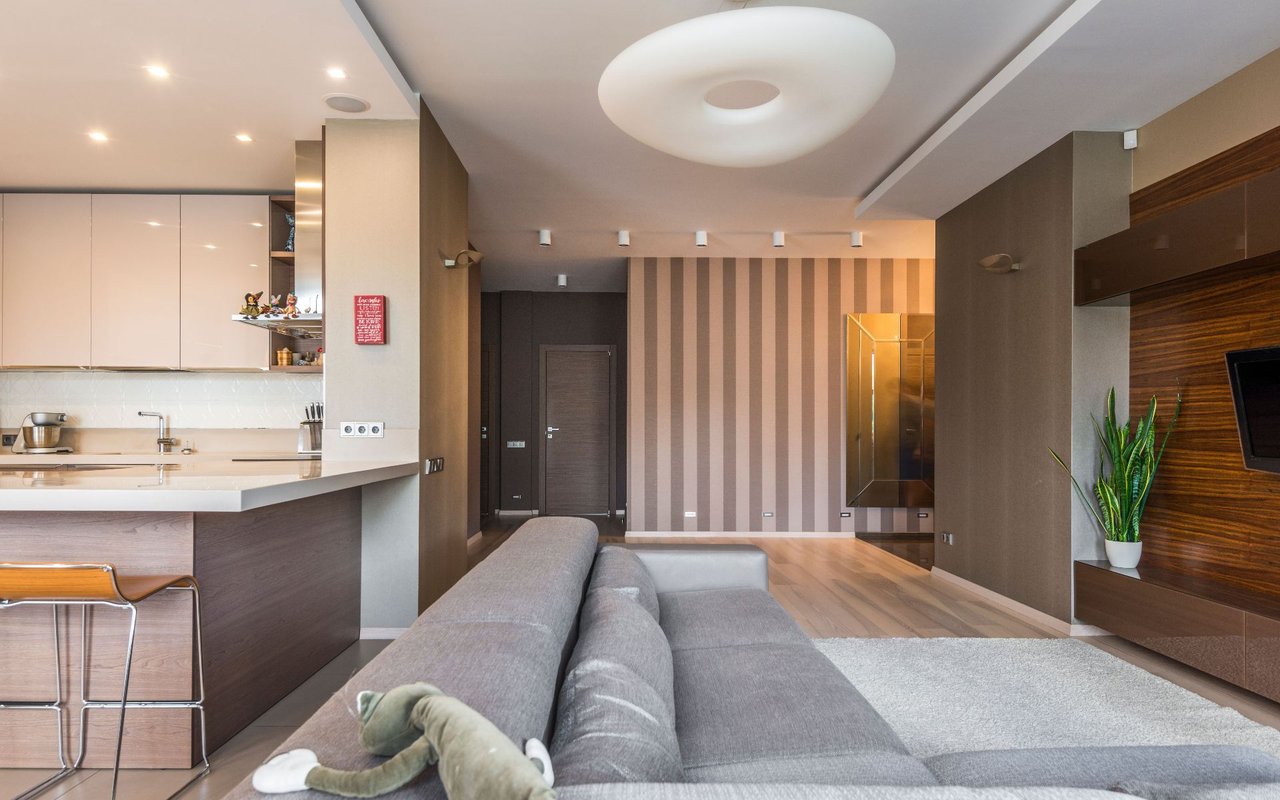 Open-plan living room with gray sectional, white kitchen, striped accent wall, and plush toys on the sofa.