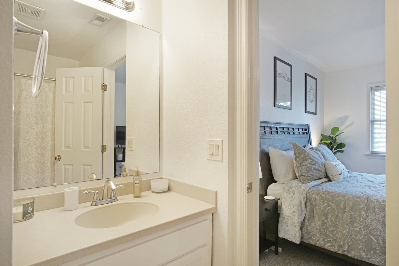 A bright bathroom with a white countertop and a sink is adjacent to a cozy bedroom. The bedroom features a bed with gray bedding, pillows, and wall art.