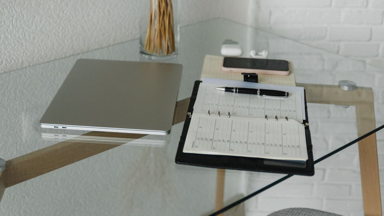 Minimal workspace with a closed laptop, open planner and pen on a glass desk