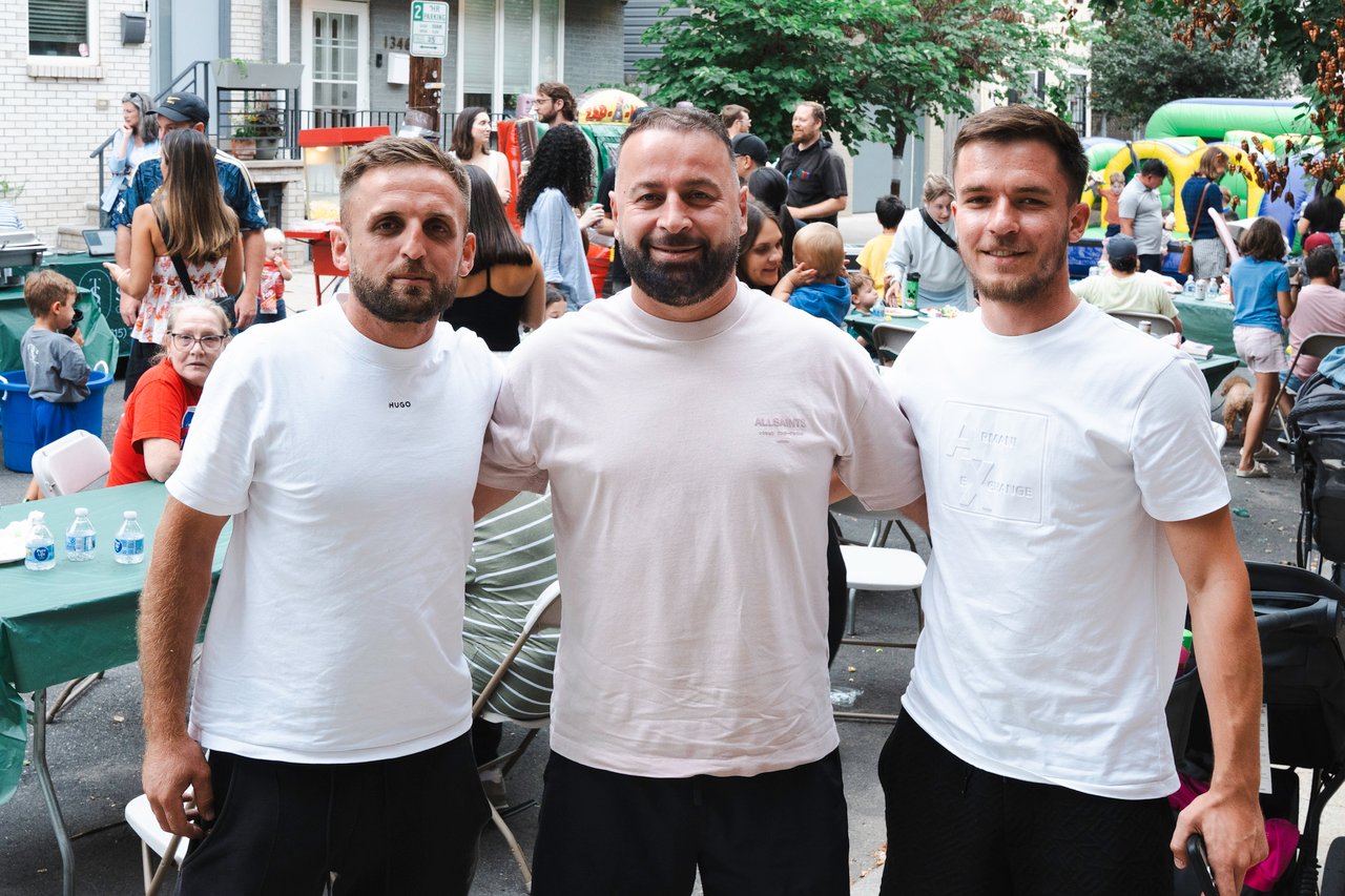 Three men in white T-shirts pose at a neighborhood block party with a crowd and inflatable play structure behind them.