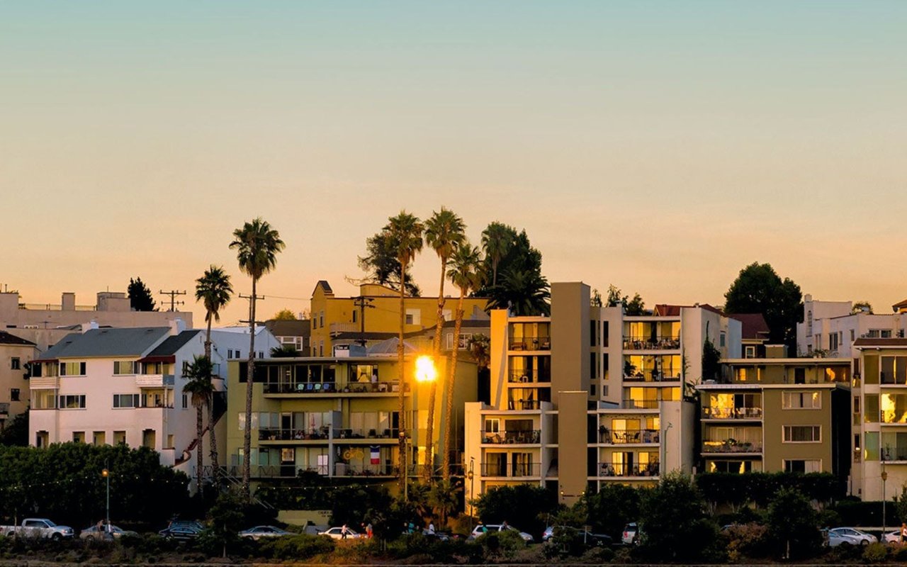 Apartment buildings and palm trees glow in the setting sun above a city street.