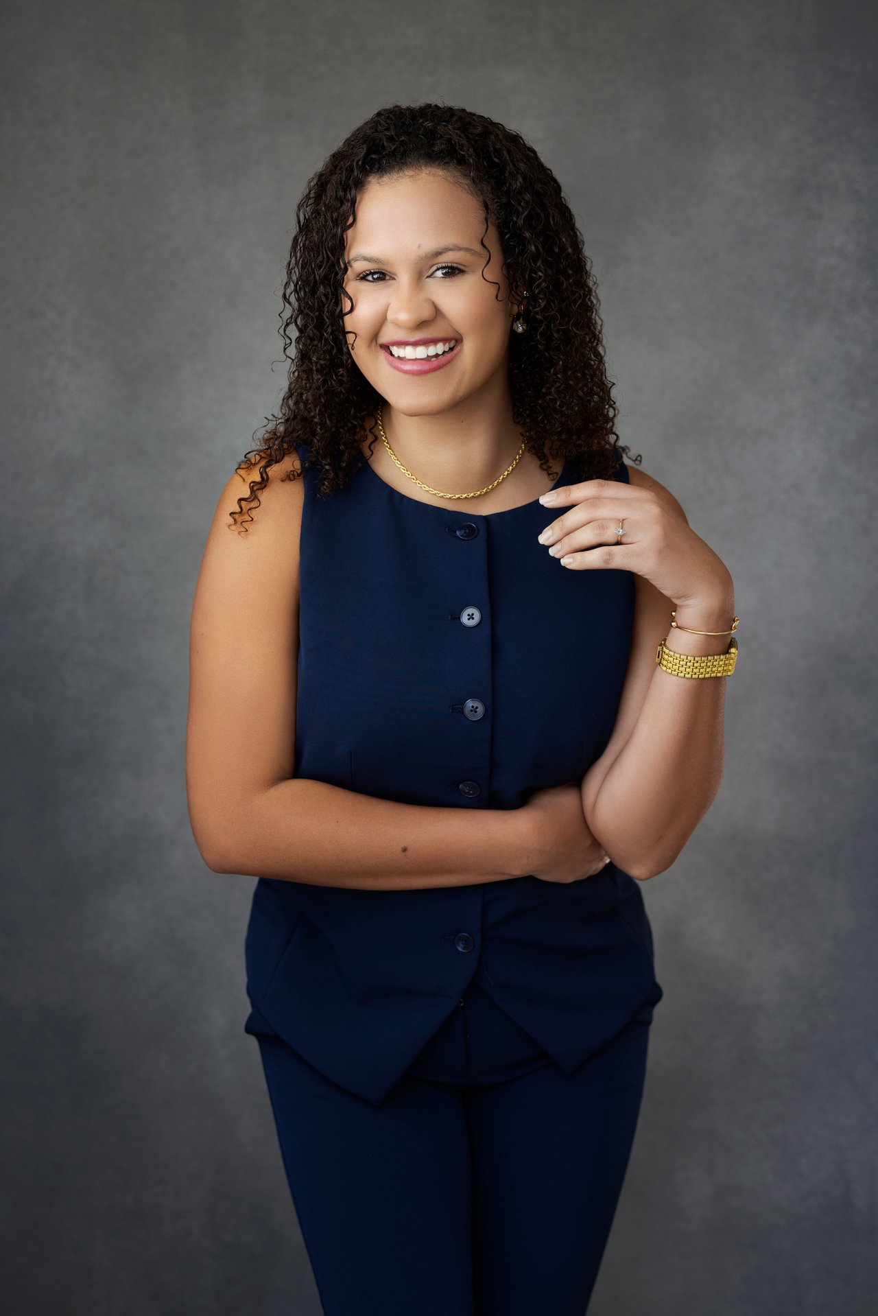 Carolyne De Souza smiling in a navy suit against a gray studio backdrop.