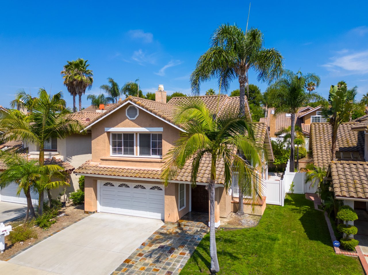 Two-story tan stucco home with a tile roof, stone driveway, and palm trees.