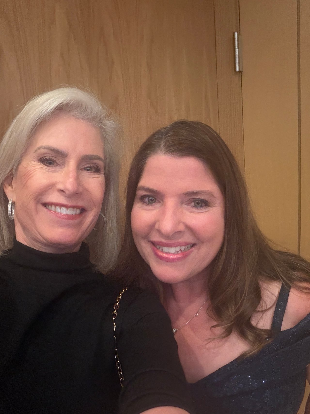 Two smiling women pose for a selfie in front of a wood-paneled wall.
