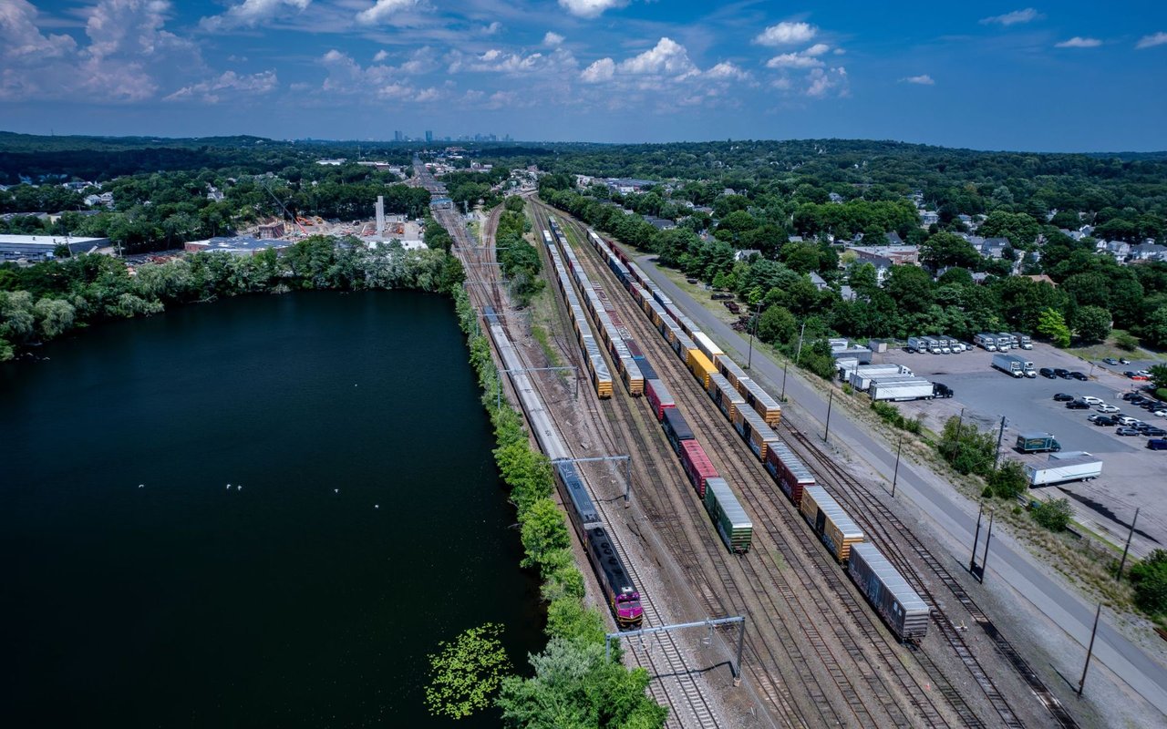 Aerial view of freight trains beside a pond, with wooded neighborhoods and a distant skyline.