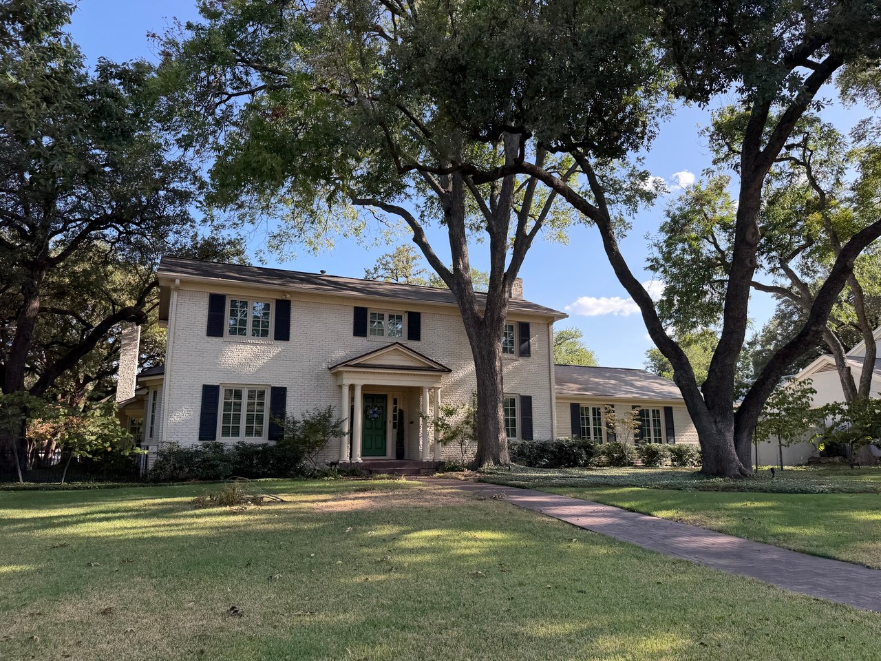 White brick two-story house with a green front door, shaded by mature trees.