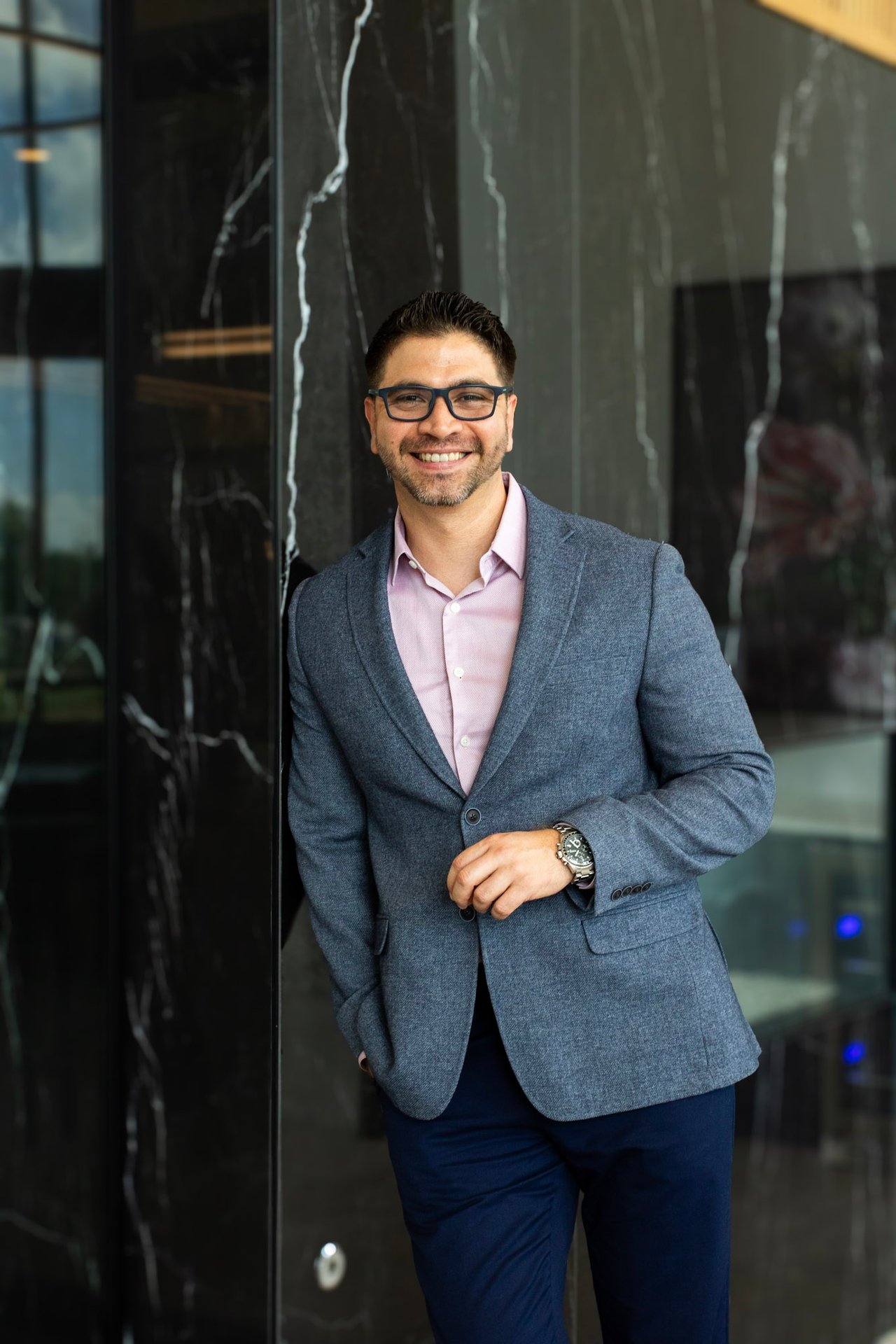Reimy Chavez Perche wearing glasses and a blue-gray blazer beside a dark stone facade