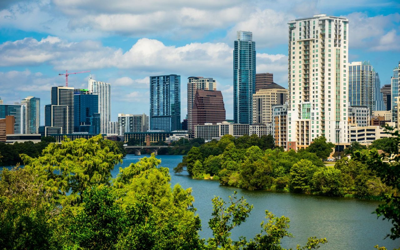 Austin downtown skyline rises above the Colorado River, framed by green trees.