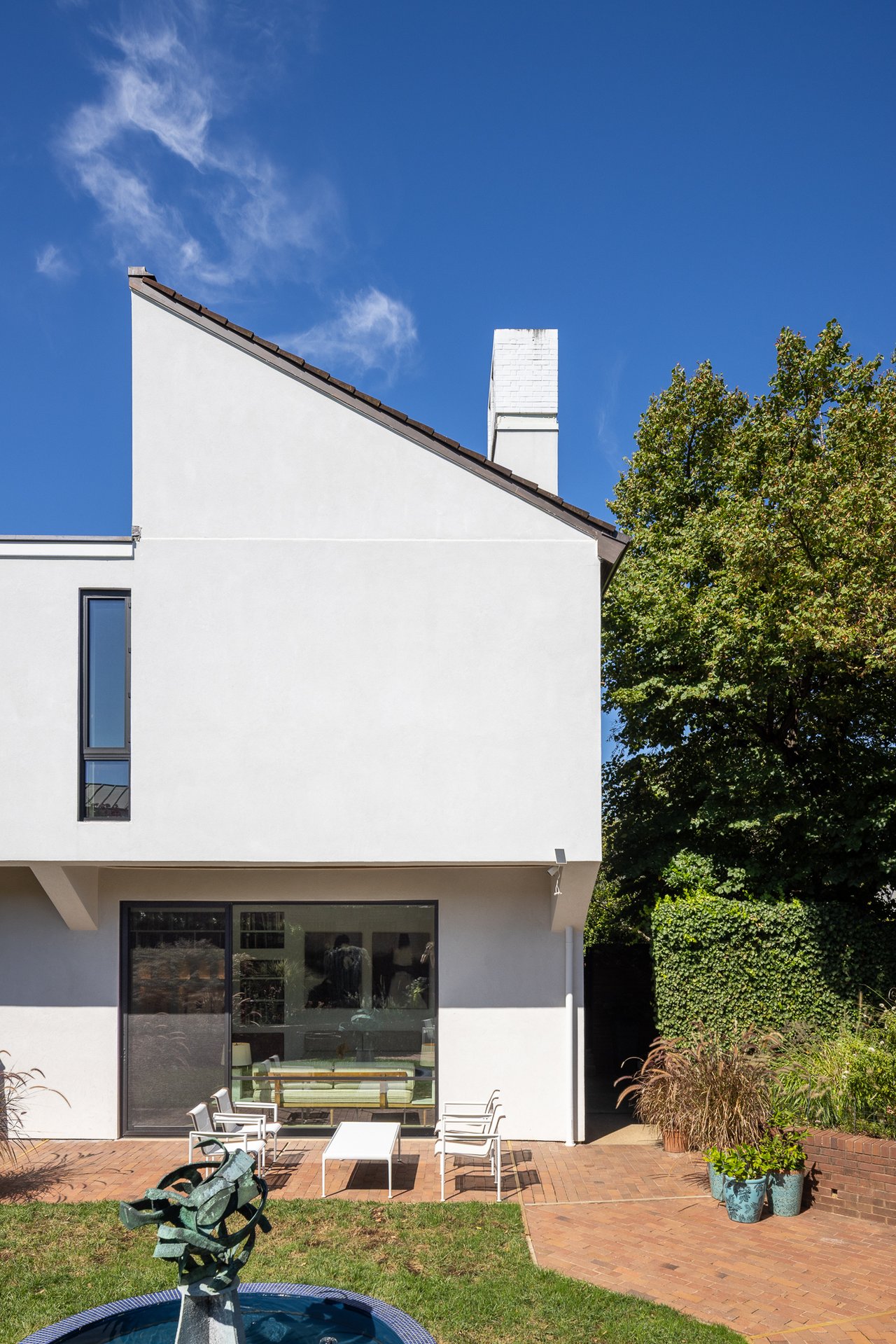 Modern white house with a sloped roof, brick patio, outdoor seating, and garden fountain.