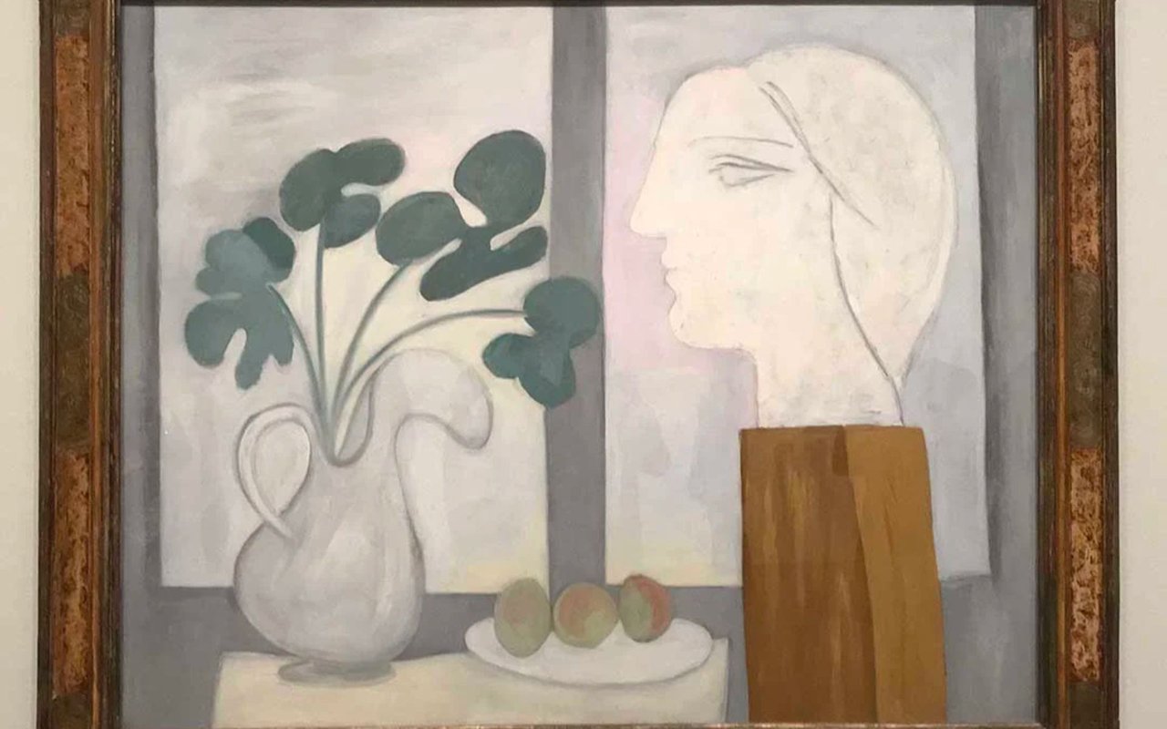 Framed painting of a woman’s profile beside a vase of flowers and fruit.