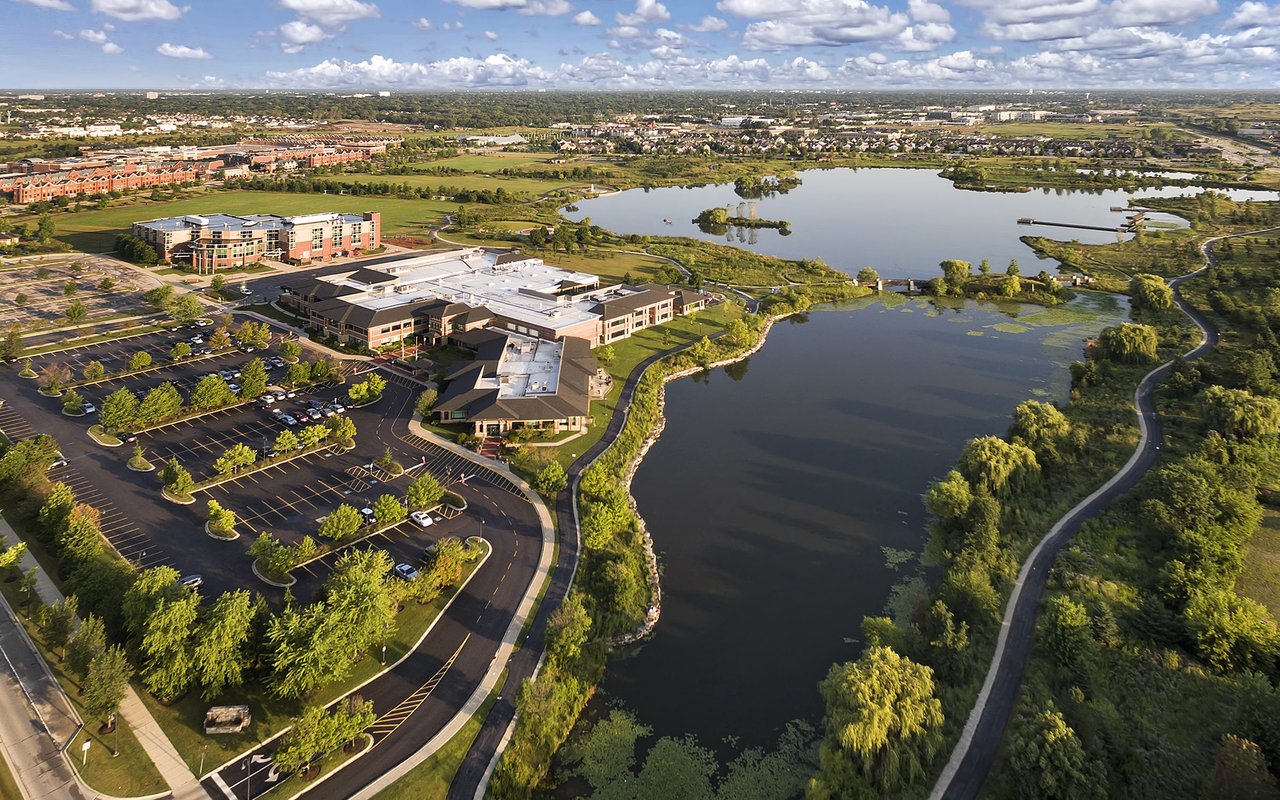 Aerial view of a commercial complex beside a lake, with tree-lined paths and parking lots.