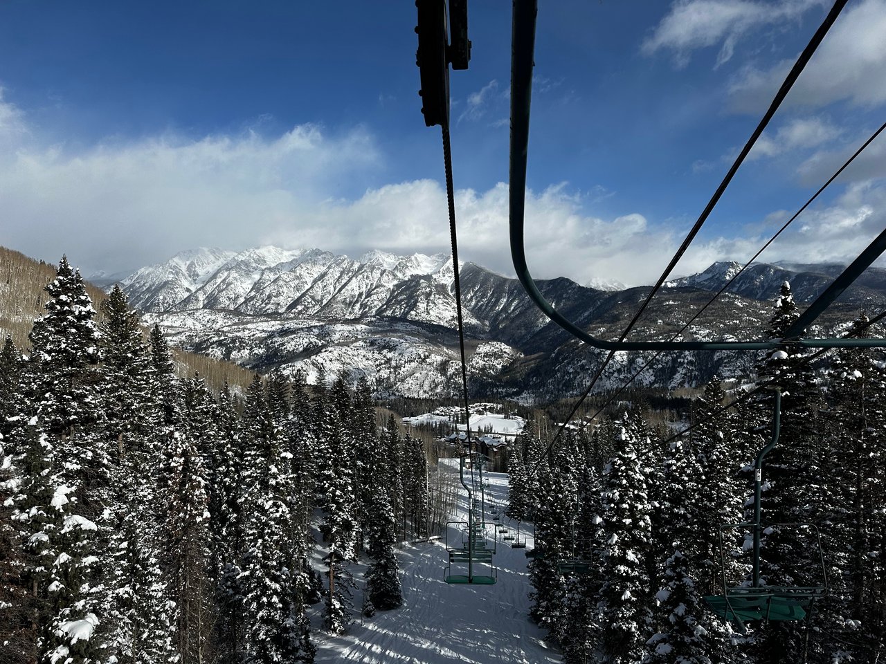 Ski lift above snow-covered evergreens, with a wide range of snowy mountains in the distance.